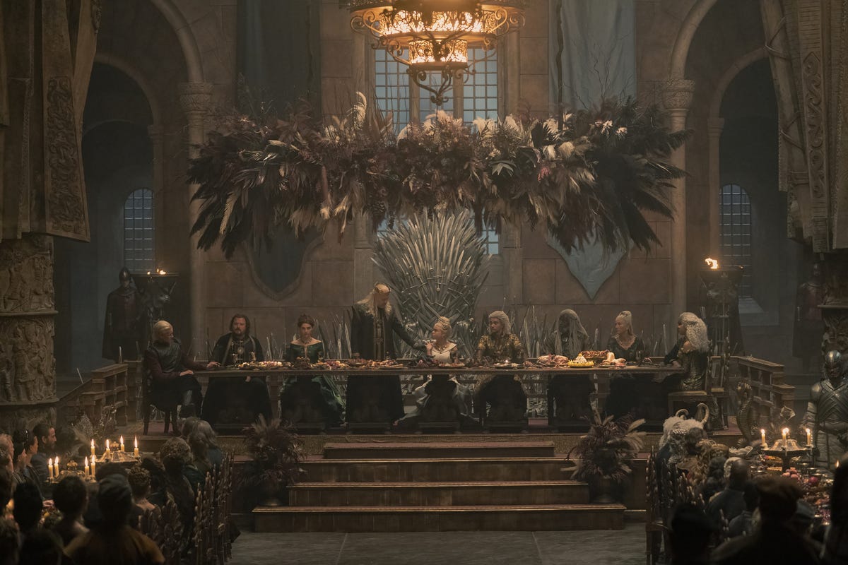 A long banquet table in a gloomy castle hall, with nine people sitting side by side